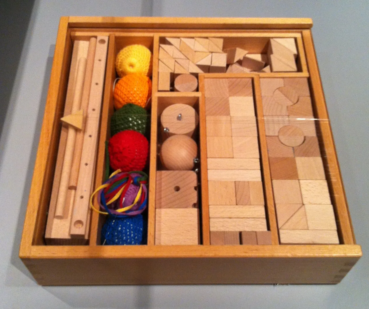 Los regalos de Fröbel Caja de madera abierta con piezas geométricas de madera clara: cubos, prismas y tablillas ordenados en compartimentos, sobre fondo oscuro