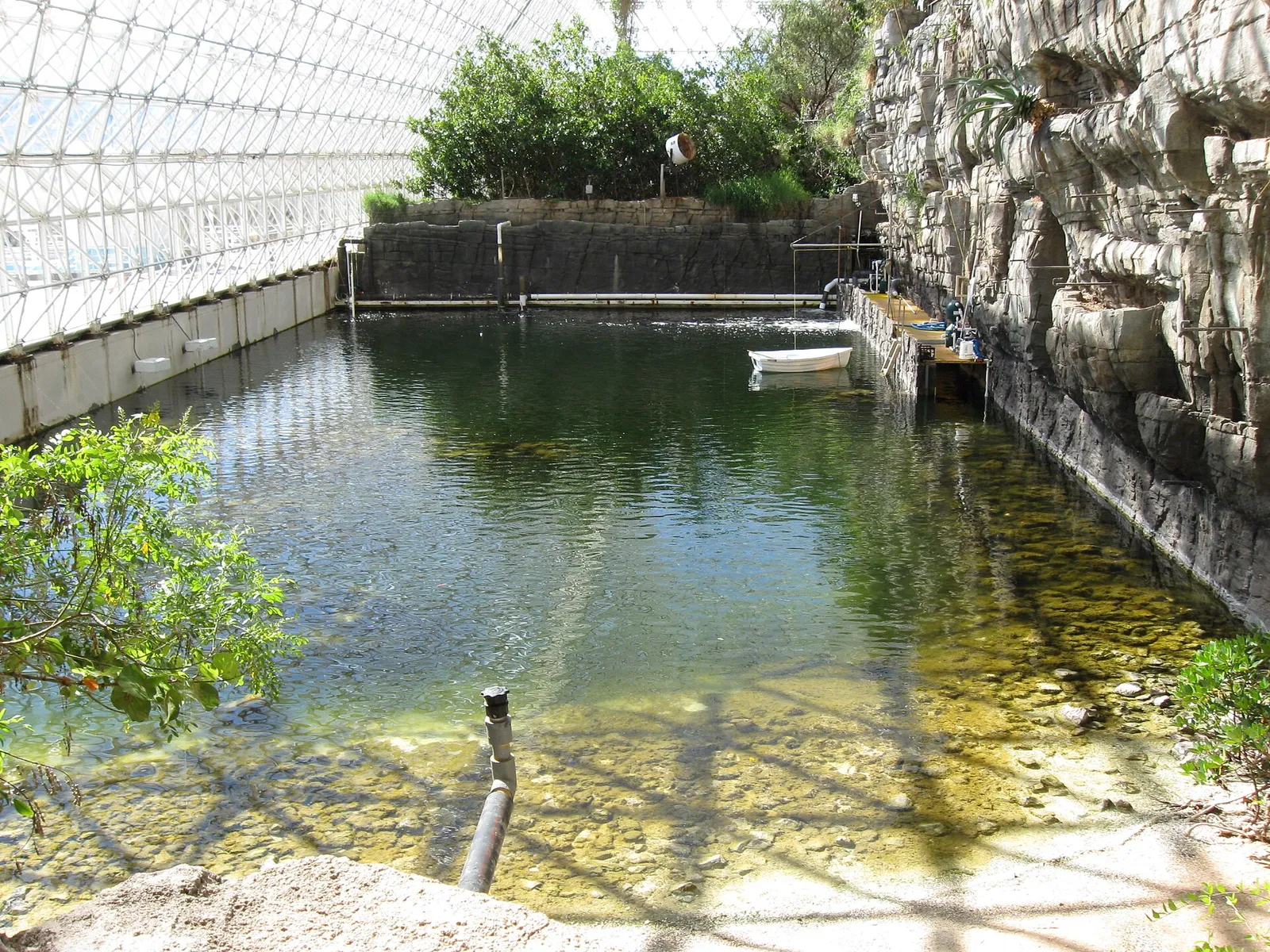 El océano de Biosfera 2 Estanque de agua clara dentro de Biosfera 2, bajo la estructura de vidrio, con una pared de roca artificial a la derecha y una pequeña lancha al fondo