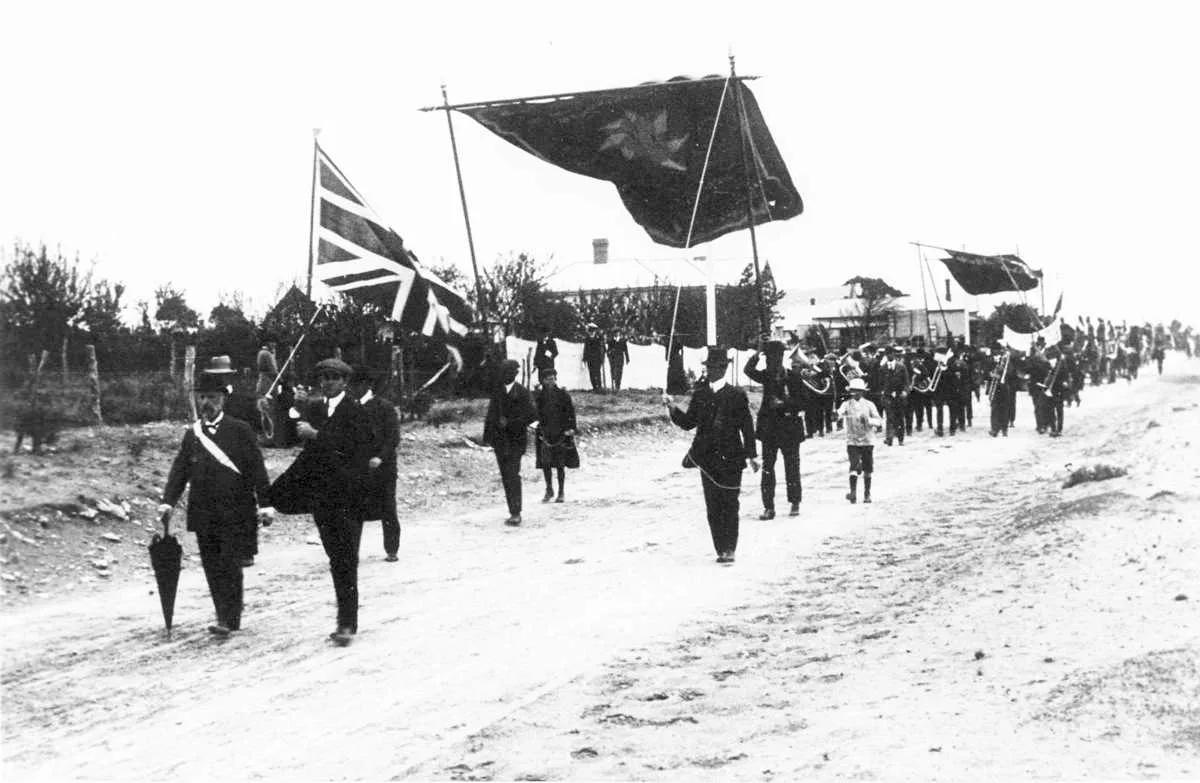 Procesión del Día de las Ocho Horas, 1912 Fotografía antigua en sepia de una procesión: hombres con sombrero marchando por una calle de tierra detrás de grandes estandartes de tela