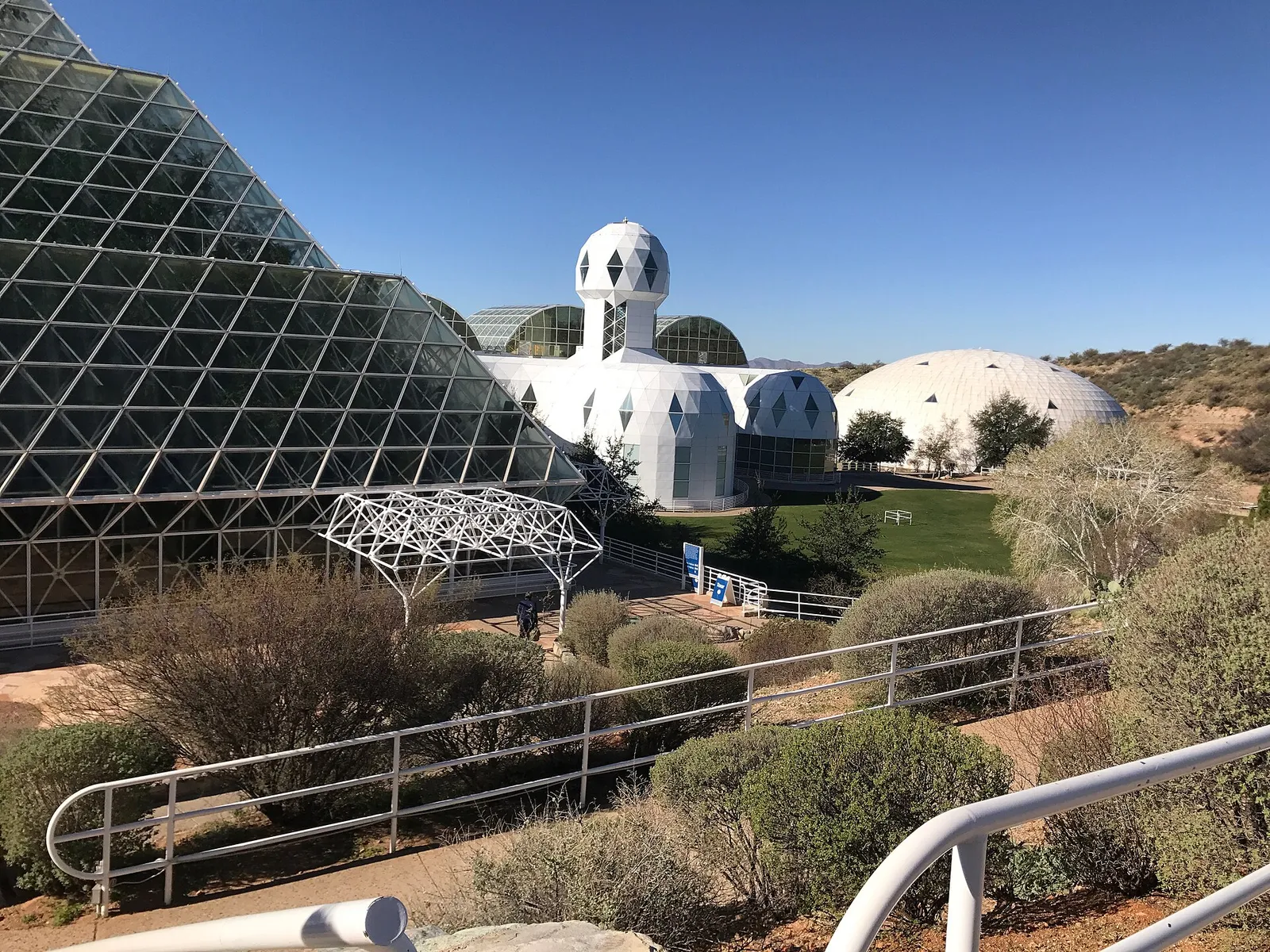 Exterior de Biosfera 2: estructuras de vidrio y acero junto a cúpulas blancas y, al fondo, el domo de uno de sus pulmones, en el desierto bajo un cielo despejado