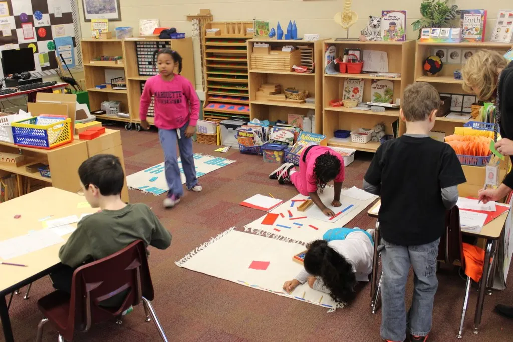 Salón Montessori Salón de clases Montessori: una niña trabaja en el piso sobre un tapete con piezas de colores, otros niños caminan entre mesas y estantes con materiales